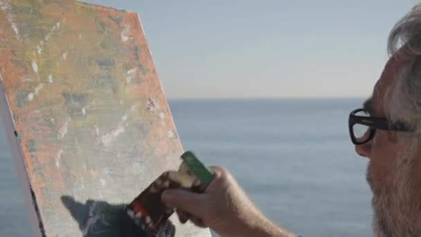 Un hombre mayor pinta un cuadro en la playa. Anciano artista masculino pintando cuadro abstracto moderno con espátula sobre fondo de mar tranquilo por la mañana. Primer plano . — Vídeos de Stock