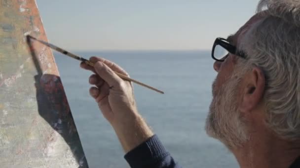 Un aîné peint un tableau sur la plage. Artiste masculin âgé peignant image abstraite moderne avec pinceau sur fond de mer calme du matin. Gros plan . — Video