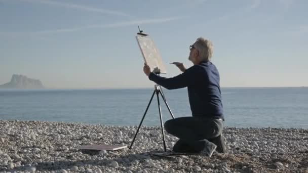 Un hombre mayor pinta un cuadro en la playa. Vista posterior del anciano artista masculino hace los últimos trazos en el lienzo con espátula ancha en la playa del mar de la salida del sol del guijarro . — Vídeo de stock