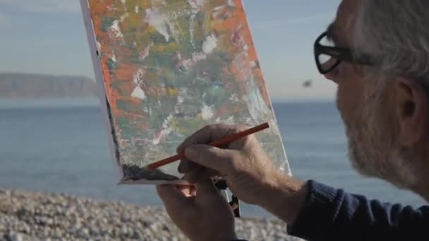 Un hombre mayor pinta un cuadro en la playa. Anciano artista masculino firma su imagen abstracta moderna en el fondo del mar calma mañana. Primer plano . — Vídeo de stock