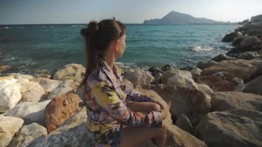 Günbatımı dağlar ve deniz kıyısında arka plan üzerinde mesafe bakarak taş üzerinde oturan genç çekici kadın