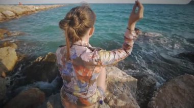 Attractive caucasian woman throws pebbles into the sea sitting on rock stone on the sunset sea coast background. Rear angle shot.