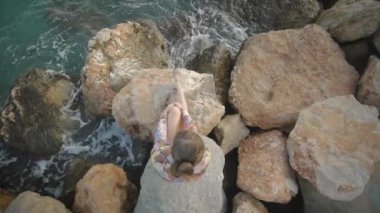 Seyir vasıl günbatımı güneş bir kayalık deniz kıyısında oturan genç çekici kadın. Rüzgarda çırpınan saç. Yüksek açısı shot.