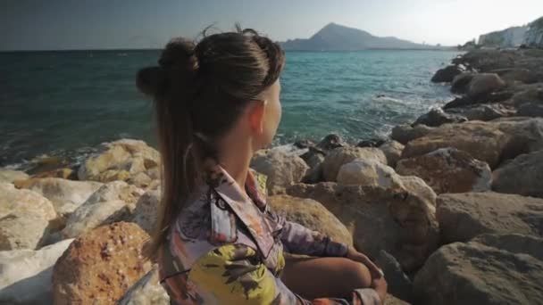 Young attractive woman sitting on rock stone looking into the distance on the sunset mountains and sea coast background. — Stockvideo