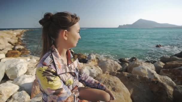 Junge attraktive Frau lächelnd auf Felsen sitzend auf den Bergen des Sonnenuntergangs und dem Hintergrund der Meeresküste. das Haar flattert im Wind. — Stockvideo