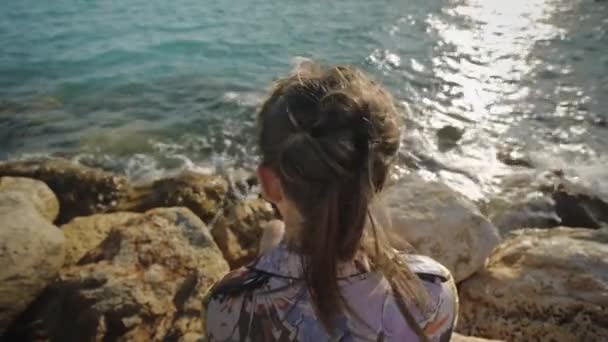 Giovane donna attraente guardando il sole del tramonto seduto su una riva rocciosa del mare. I capelli svolazzano nel vento. Colpo angolo posteriore . — Video Stock
