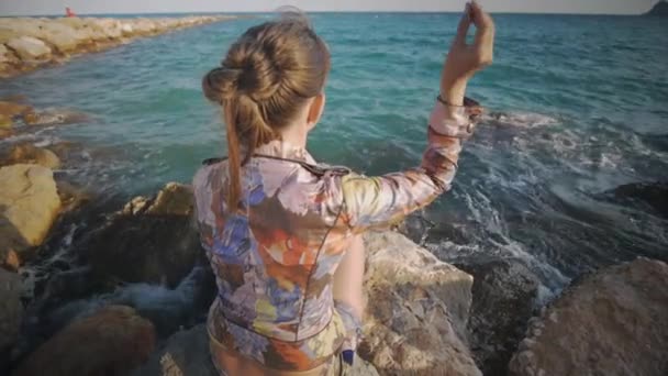 Attractive caucasian woman throws pebbles into the sea sitting on rock stone on the sunset sea coast background. Rear angle shot. — Stok video