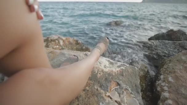 若い魅力的な女性の夕日岩の海の海岸に座って見て。風になびく髪。ドリーは撃たれた. — ストック動画