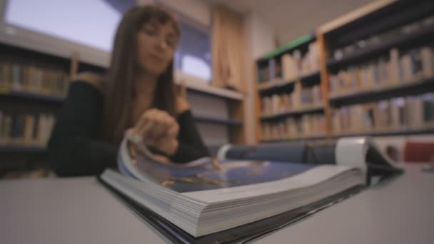 Jeune femme caucasienne tourne la page du livre illustré dans la bibliothèque. Prise de vue déconcentrée . — Video