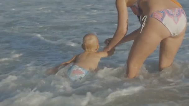 Young caucasian mother playing with two years old blonde baby enjoying water waves splashes foam holding hand on sea beach. Happy family concept. — 비디오