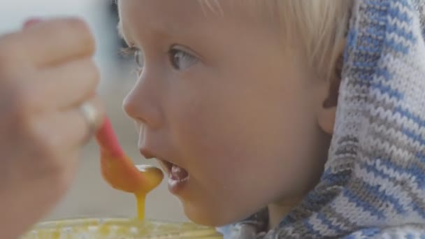Nahaufnahme der weiblichen Hand Fütterung mit einem Löffel blondes Baby in gestricktem Kapuzenpullover am Meer Hintergrund. — Stockvideo