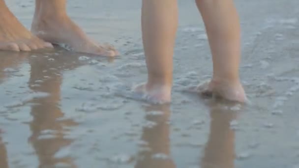 Mujer joven caucásica y bebé de dos años jugando en la playa del atardecer. Primer plano de las piernas de mamá e hija cerca del mar dejando huellas en la arena mojada . — Vídeo de stock