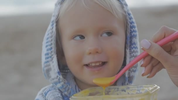 Närbild av kvinnlig hand utfodring med en sked blond baby i stickad hoodie på seaside bakgrund. — Stockvideo