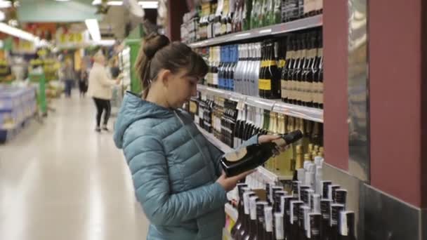 Mujer en el supermercado. Mujer joven caucásica en chaqueta azul eligiendo vino espumoso en los estantes de la tienda fondo . — Vídeos de Stock