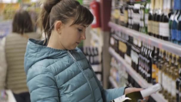 Mujer en el supermercado. Joven mujer caucásica en chaqueta azul lee la etiqueta en la pequeña botella eligiendo vino espumoso . — Vídeos de Stock