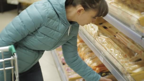 Kobieta w supermarkecie. Widok z tyłu z młodych kaukaski kobieta w Błękitnej Kurtki wybierając bagietki chleb, wprowadzenie go w koszyku. — Wideo stockowe