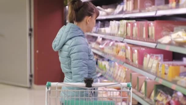 Frau im Supermarkt. junge kaukasische Frau in blauer Jacke liest Etikett von Käse und legt es in den Einkaufswagen. — Stockvideo