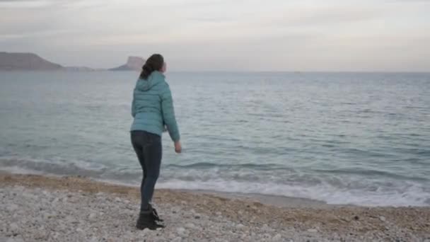 在孤独的寒冷海滩的女人。高加索妇女向平静的大海投掷石块. — 图库视频影像