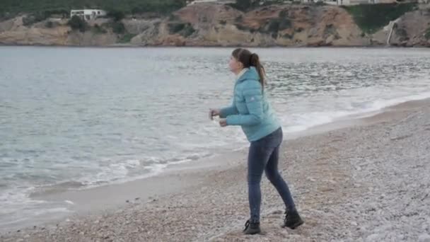 Frau am einsamen kalten Strand. Kaukasierin wirft Steine ins ruhige Meer. — Stockvideo