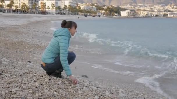 Mujer en la playa fría y solitaria. Mujer caucásica rebota de la ola que viene jugando con piedras cerca del agua en la playa . — Vídeos de Stock