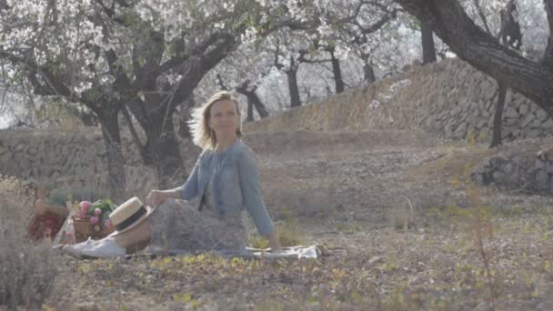 Heureuse femme caucasienne souriante s'amusant assis sur le sol à l'extérieur sur pique-nique sur fond d'amandiers en fleurs . — Video