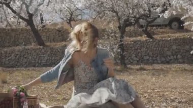 Çiçek Bahçe kadında. Tartan ekose piknik gıda ile üzerinde oturan seksi beyaz kadın. Rüzgar Bora ağaçlar çiçek alır ve masadan bir şey alır.