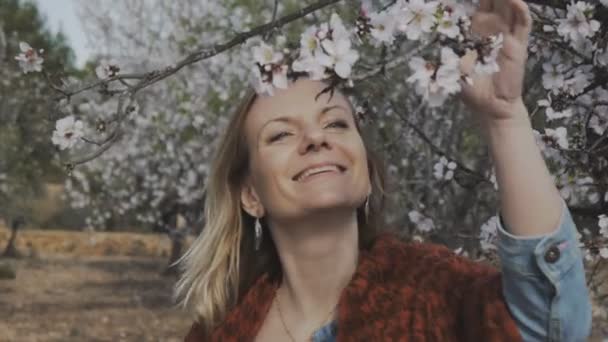 Mujer en el jardín floreciente. Primer plano de la atractiva mujer rubia sonriente disfrutando del olor a flor de almendra en el parque de primavera . — Vídeo de stock