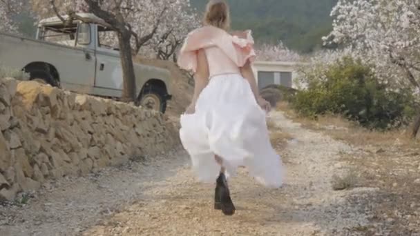 Mujer en el jardín floreciente. Vista trasera de mujer rubia caucásica vestida en estilo ecléctico caminando por el camino de tierra a la camioneta abandonada. Movimiento lento . — Vídeo de stock