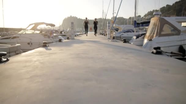 Young women doing exercises at sea port. Fit attractive caucasian girls run along the pier between the yachts. — Stock Video