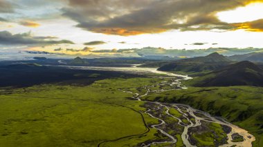 Yeşil yosun arazisinden akan İzlanda 'nın örülmüş nehirlerinin hava perspektifi volkanik tepeler, uzak buzullar ve sıcak turuncu bir gökyüzü..