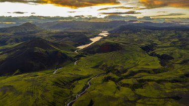 Yansıtıcı bir göl, kıvrımlı nehirler, yeşil tepeler ve turuncu ve sarı renklerle aydınlatılmış gökyüzü içeren İzlanda 'nın volkanik arazisinin havadan görüntüsü..