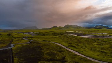 İzlanda 'nın yosunlarının hava perspektifi ovaları, kıvrımlı nehirleri ve fırtınalı bulutlar ve sıcak güneş ışığıyla dinamik bir gökyüzü altında engebeli dağları kaplıyordu..