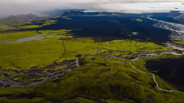 İzlanda 'nın yosun örtülü tepeleri, karanlık volkanik düzlükleri, örülmüş nehirleri ve bulutlu bir gökyüzünün altında bir buzulun havadan bakış açısı..