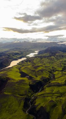 İzlanda 'nın yeşil yosunlarının hava perspektifi tepeleri, karanlık volkanik toprağı, dolambaçlı bir nehir, yansıtıcı bir göl ve uzak sıradağlar..
