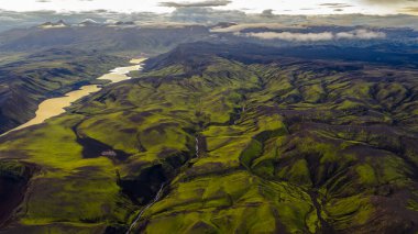 İzlanda 'nın yeşil tepeleri, volkanik arazisi, dolambaçlı yansıtıcı nehri ve bulutlu bir gökyüzünün altında engebeli dağları olan havadan bakış açısı..