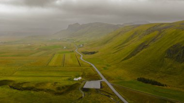 İzlanda 'nın kırsal kesimindeki yosun kaplı tepelerle çevrili yeşil alanların havadan perspektifi. Dolambaçlı bir yol, bulutlu bir gökyüzünün altındaki dağınık çiftlik evlerine götürür..