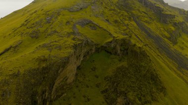 İzlanda 'daki engebeli bir volkanik yamacın havadan perspektifi. Dik uçurumlar, derin yarıklar, koyu kaya oluşumları ve canlı yeşil yosunlar..