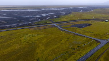 İzlanda 'daki yosunlu arazide dönen bir yolun hava görüntüsü, örülmüş nehirlerle çevrili siyah kum düzlükleriyle çevrili. Yeşil çatılı bir bina görünüyor..