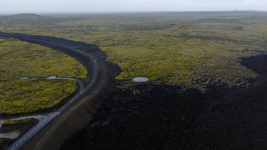 Karanlık lav tarlaları, yeşil yosun yamaları ve bulutlu bir gökyüzü altında dolambaçlı bir yol içeren İzlanda 'nın volkanik arazisinin hava görüntüsü..