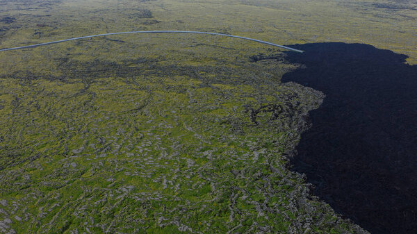An aerial perspective of Iceland's green moss covered landscape meeting a dark volcanic field, with a curving road cutting through the scene.