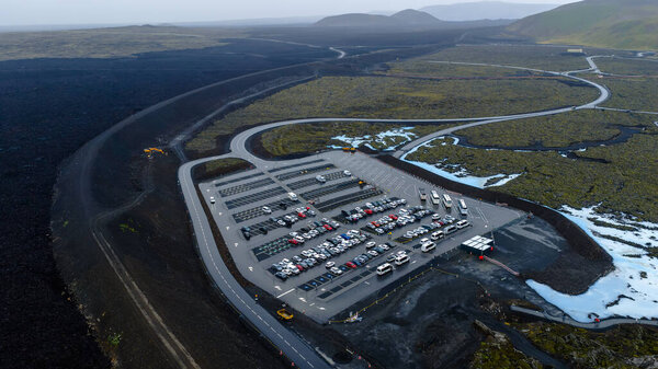 Воздушное изображение парковки с автомобилями и автобусами, окруженной черной вулканической местностью, зеленым мхом, снежными пятнами, холмами и далекими горами в Исландии.