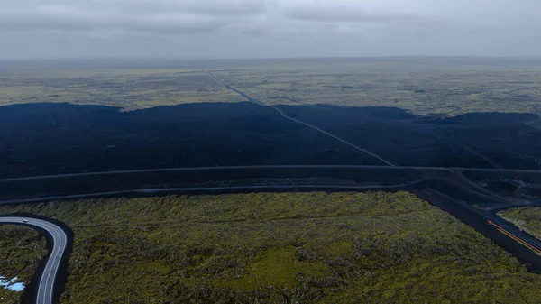 Karanlık lav tarlaları, yeşil yosunlar, dolambaçlı bir yol, bir araç ve bulutlu bir gökyüzü içeren İzlanda 'nın volkanik arazisinin hava perspektifi..
