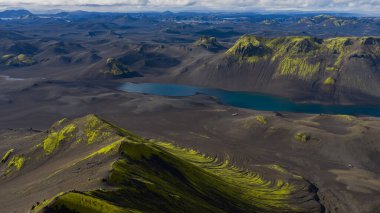 Karanlık toprak, yeşil yosun kaplı tepeler, mavi bir göl ve bulutların altındaki uzak sıradağlar içeren İzlanda 'nın volkanik arazisinin havadan perspektifi.