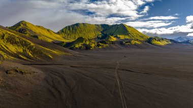 İzlanda 'da verimsiz bir volkanik arazi canlı yeşil yosun kaplı dağlar, toprak bir yol ve uzak bir atmosfer yaratan parçalı bulutlu bir gökyüzü..