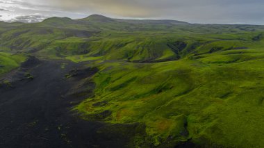 Yeşil yosun kaplı tepeler, kara toprak, çıkıntılar, vadiler ve bulutlu gökyüzünün altındaki uzak bir dağ ile İzlanda 'nın volkanik manzarasına hava perspektifi..