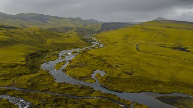 Yeşil yosunlu İzlanda 'nın volkanik arazisinin hava perspektifi, dolambaçlı bir nehir, yuvarlanan tepeler, dar bir yol ve bulutlu bir gökyüzü..