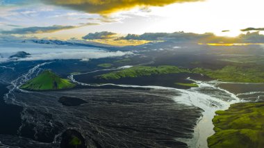 Karanlık düzlükler üzerinde yeşil bir volkanik tepenin, örülmüş nehirlerin, karla kaplı dağların ve İzlanda 'da bulutlarla parlayan bir günbatımının hava görüntüsü..