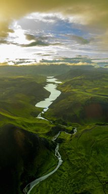 Bereketli yeşil tepeler, volkanik oluşumlar ve parçalı bulutlu gökyüzü ile çevrili, yansıtıcı göllere akan dolambaçlı bir nehrin hava perspektifi..