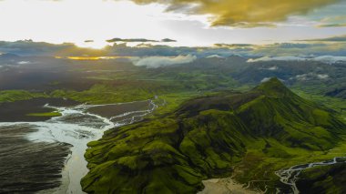 Yeşil bir volkanik dağ. Etrafı örülmüş nehirler ve altın ışığın altında siyah kum ovalarıyla çevrili. Uzak dağlar bulutlarla kısmen gizlenmiş..