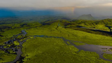 İzlanda 'nın yeşil yosunlarının hava perspektifi kıvrımlı nehirler, soluk bir gökkuşağı ve arka planda volkanik bir tepe ile kaplıydı..